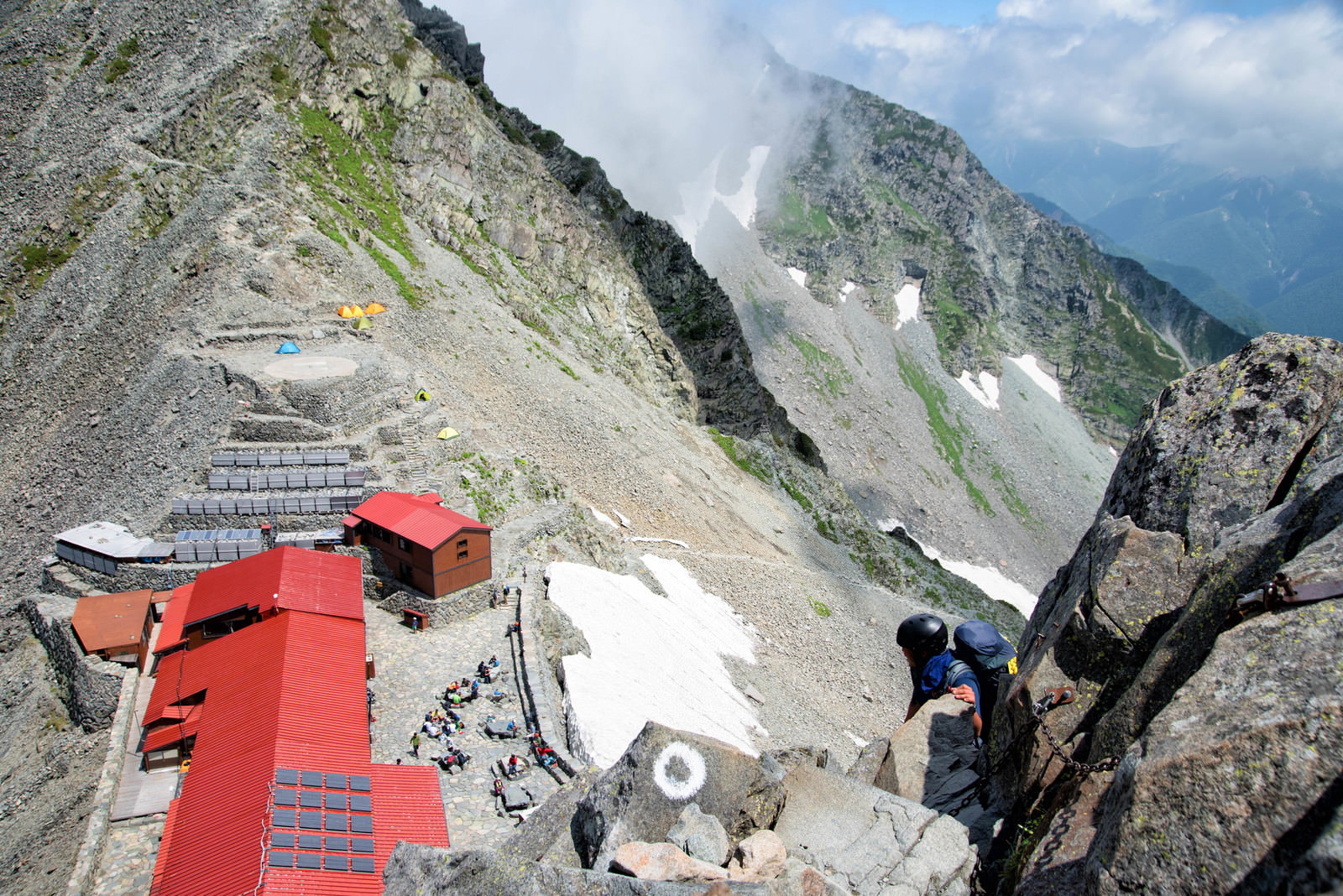 奥穂高岳の岩場にある赤い屋根の山小屋と鎖場を登る登山者
