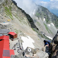 奥穂高岳の鎖場から望む赤い屋根の山小屋と登山者の写真