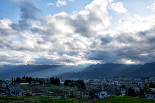 北アルプスの山並みにかかる夕暮れの光芒と住宅地の風景