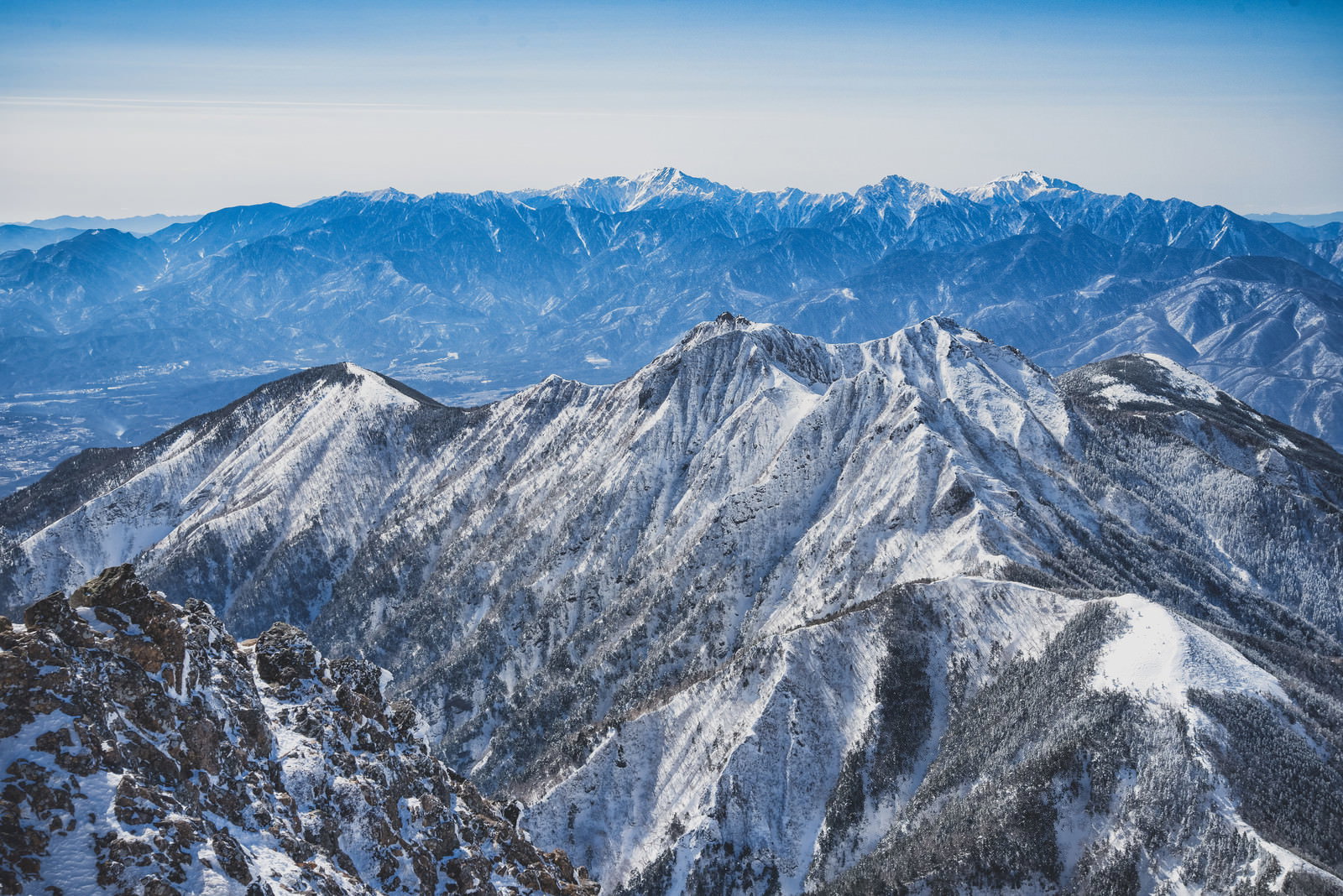 手前から奥へ連なる雪山群。白と樹林の縞状の山肌と奥の連峰が青空に映える