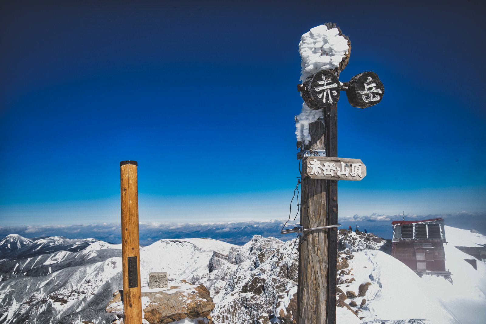雪に覆われた山頂に立つ木製の指導標と背景の雪山