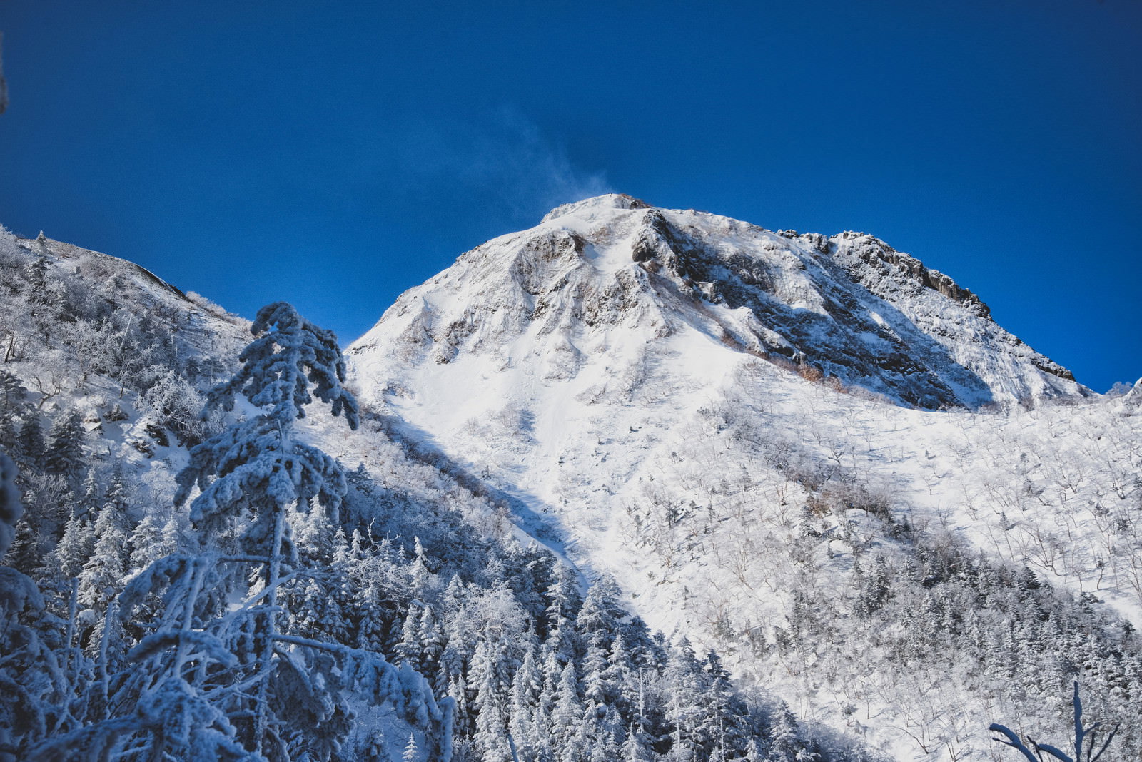 青空をバックに雪煙が舞い上がる阿弥陀岳の山頂