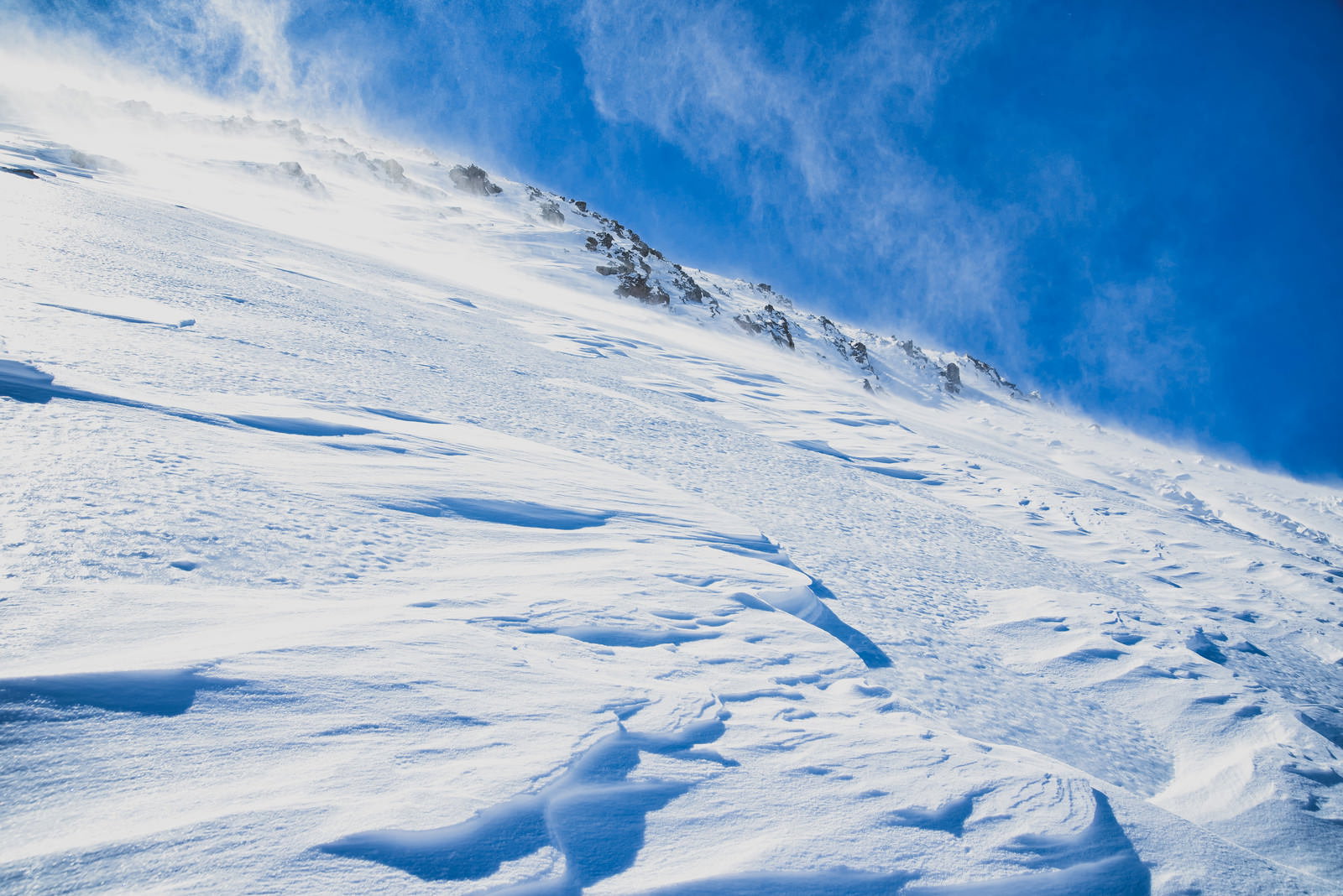 青空の下に広がる雪山の急斜面、風で雪が舞い上がる雪景色