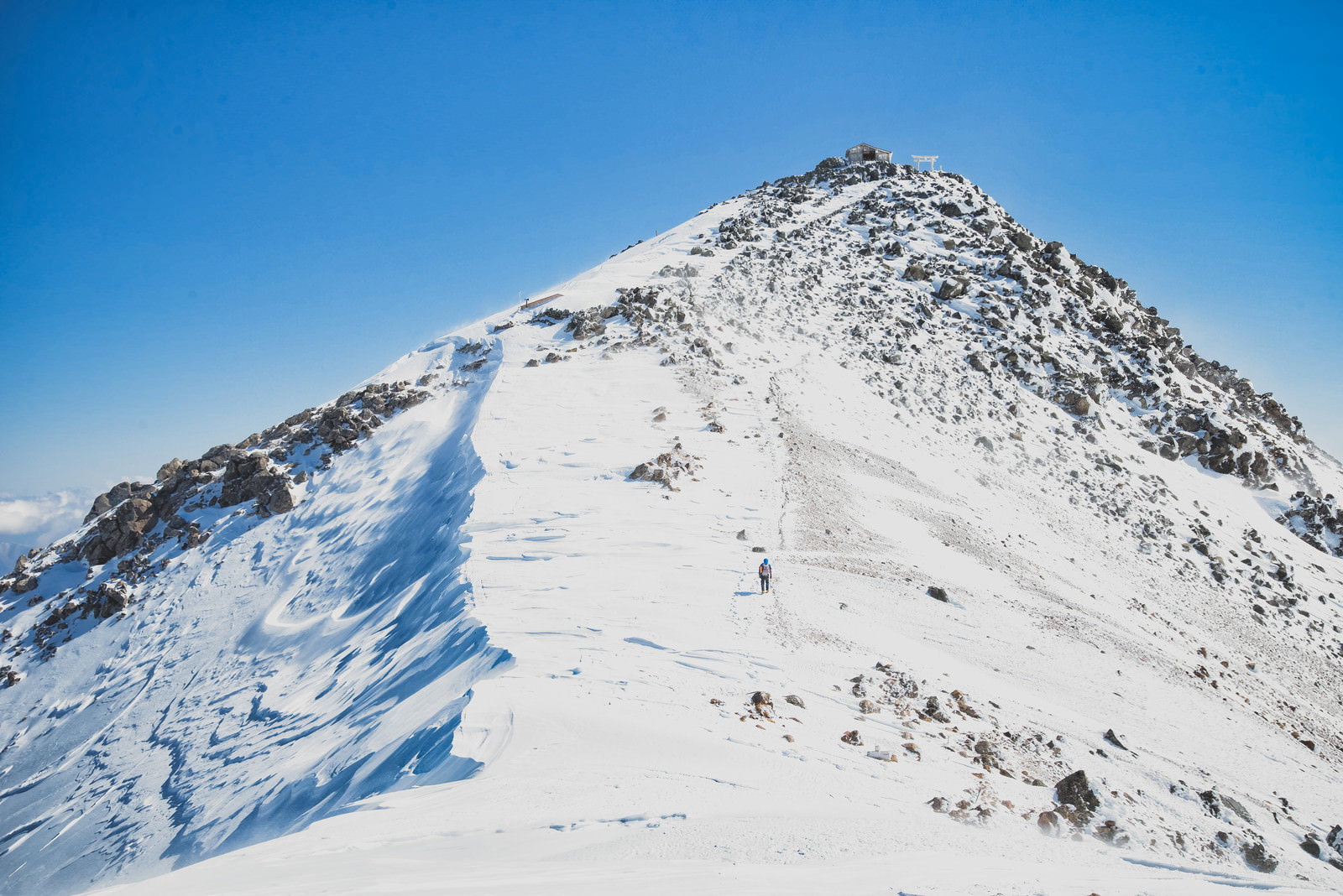 青空の下、雪に覆われた剣ヶ峰の登山道を歩く登山者