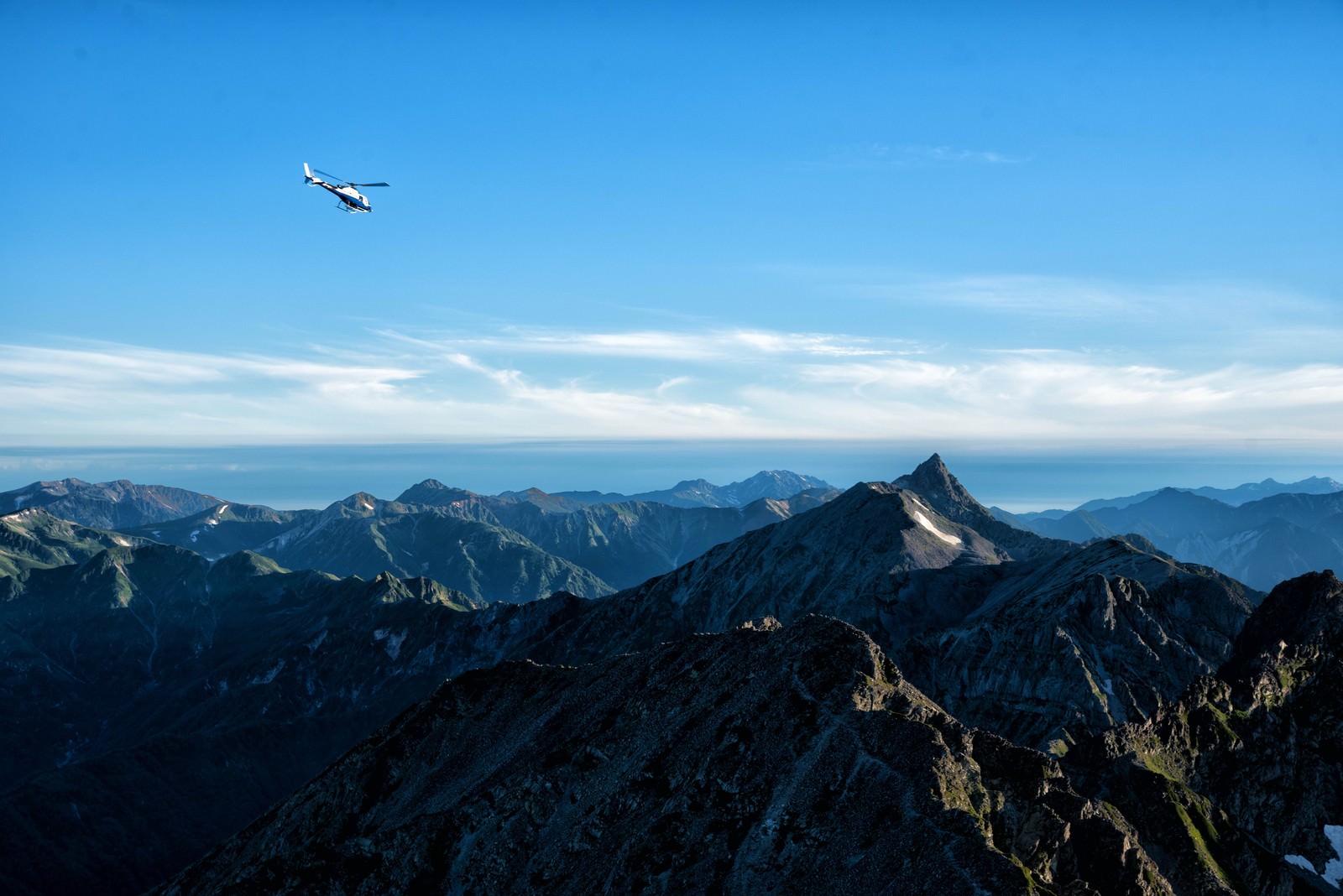青空の下、北アルプスの山々が連なる中をヘリコプターが飛ぶ風景