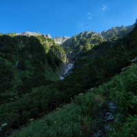 山間に見える吊尾根の岩壁と新緑の登山道を進む登山者の風景の写真