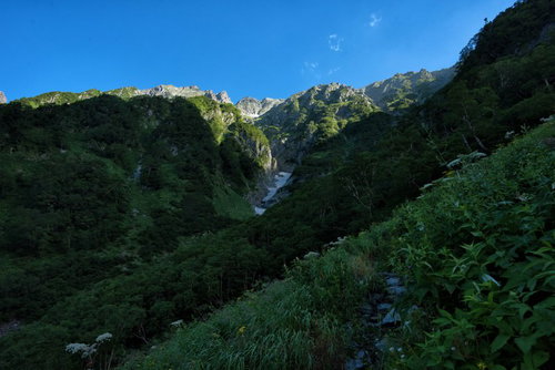山間に見える吊尾根の岩壁と新緑の登山道を進む登山者の風景
