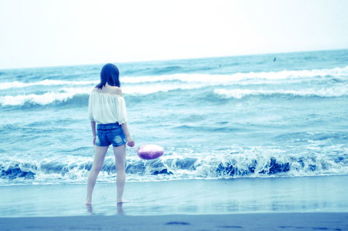 波打ち際で浅瀬の波と戯れる白いワンピース姿の若い女性の後ろ姿