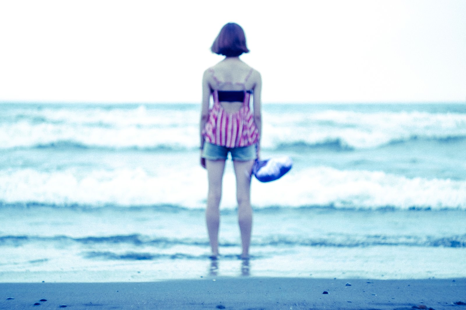 波打ち際の砂浜に立ち、青い海を見つめる女性の後ろ姿