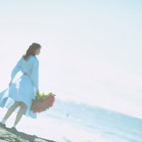 赤い花束を持って波打ち際を歩く女性の後ろ姿の写真