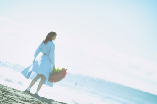 赤い花束を持って波打ち際を歩く女性の後ろ姿