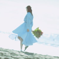 波打ち際を歩く青いスカートの女性と花束を持つ後ろ姿の写真