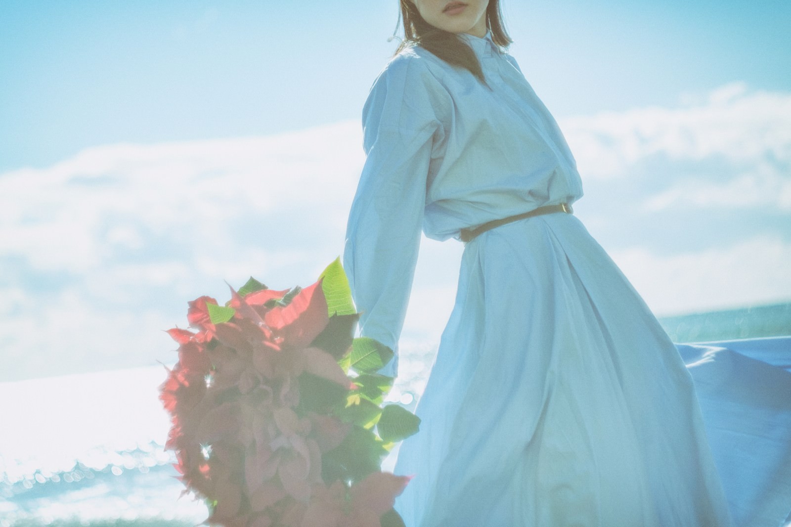 海辺で赤いポインセチアの花束を持つ水色ワンピースの女性［モデル：Mizuho］