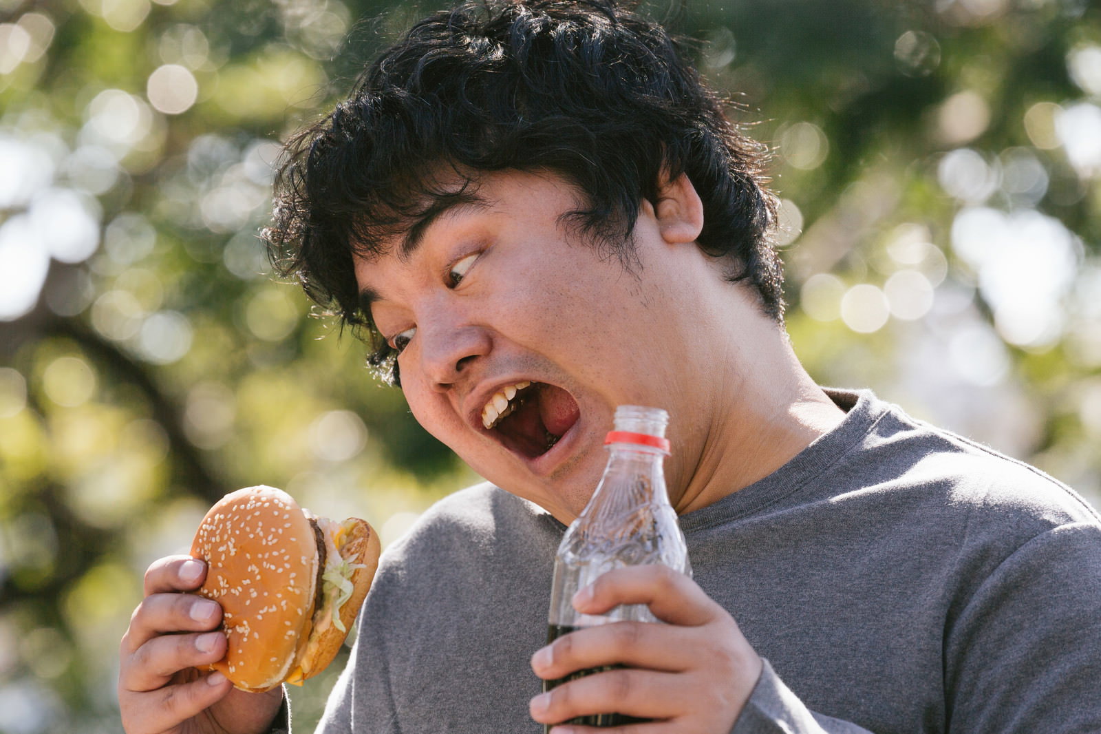 ハンバーガーと飲み物を両手に持って食べる男性［モデル：段田隼人］