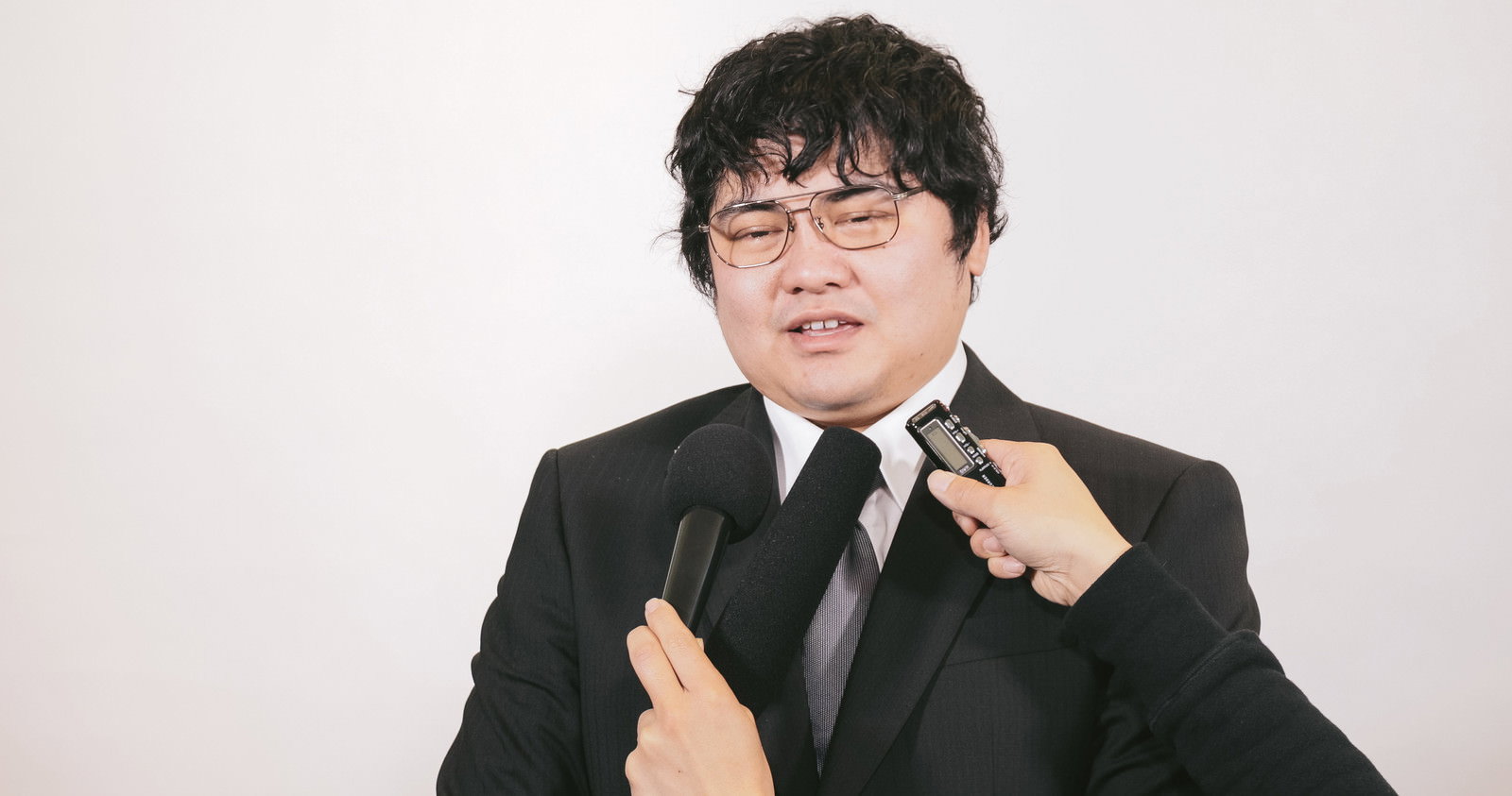 スーツを着た眼鏡の男性が、両手のマイクに向かって記者会見で応答している［モデル：段田隼人］