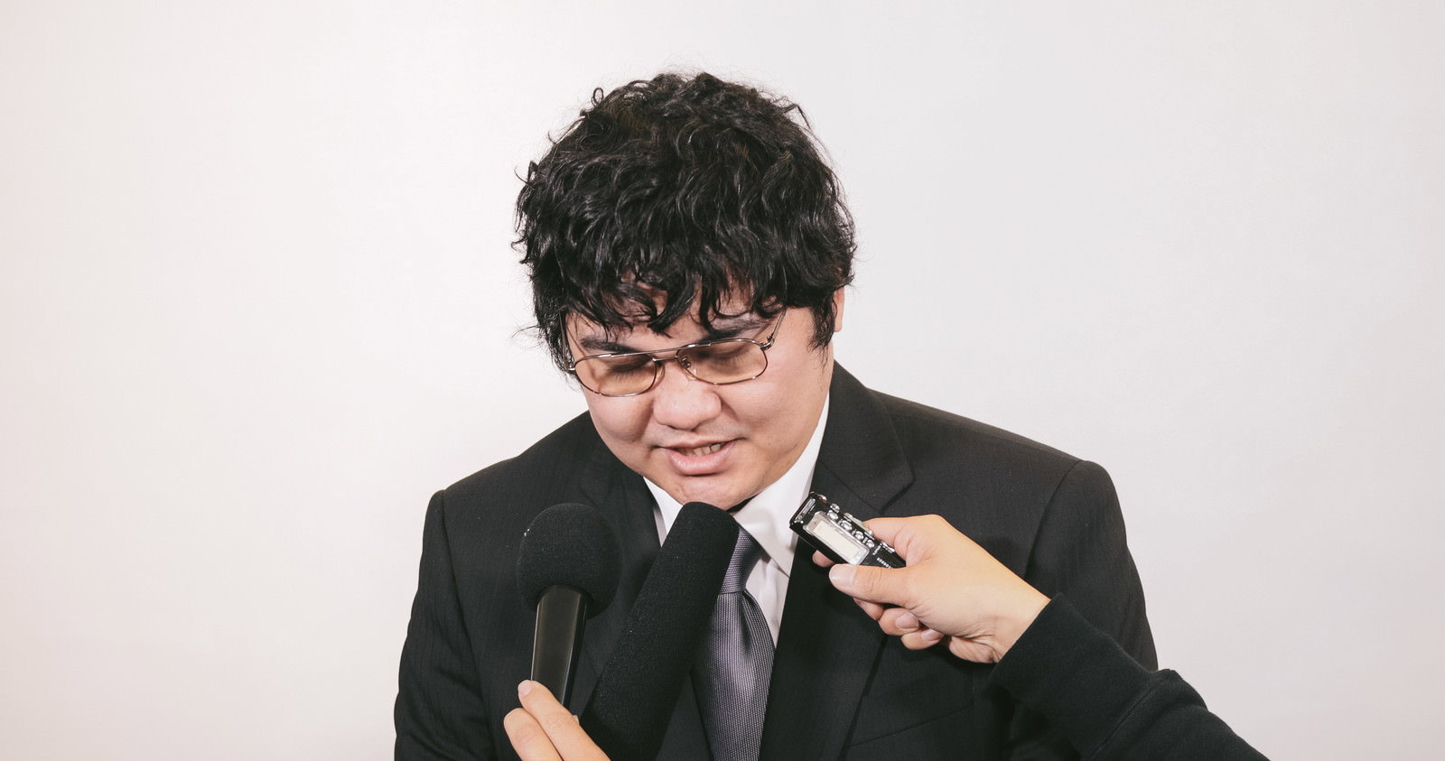 謝罪会見でマイクの前に立つ黒スーツの男性［モデル：段田隼人］