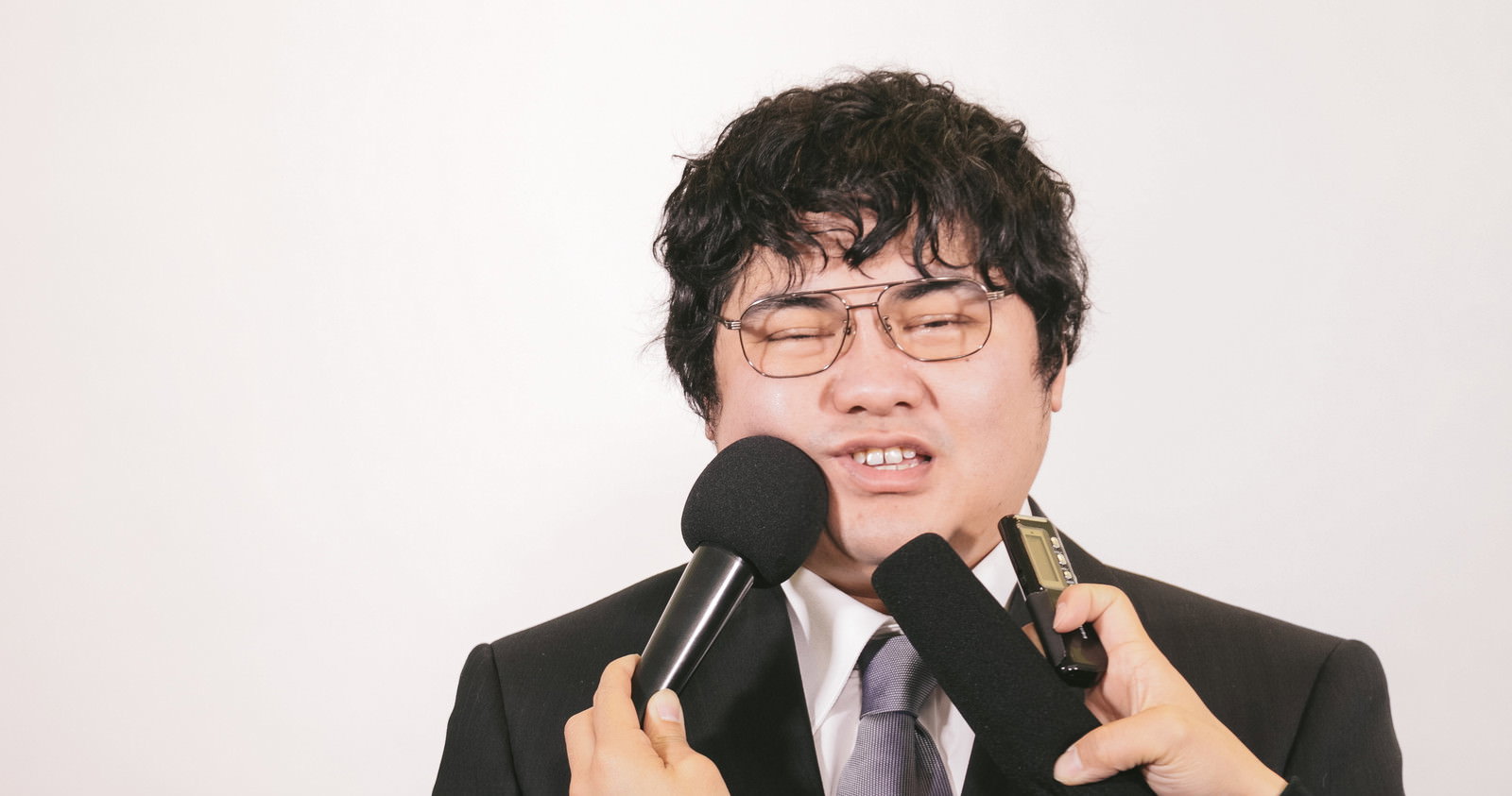 複数のマイクを向けられ困惑する眼鏡の男性［モデル：段田隼人］