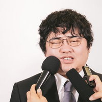 複数のマイクを向けられて困惑する男性の記者会見の写真