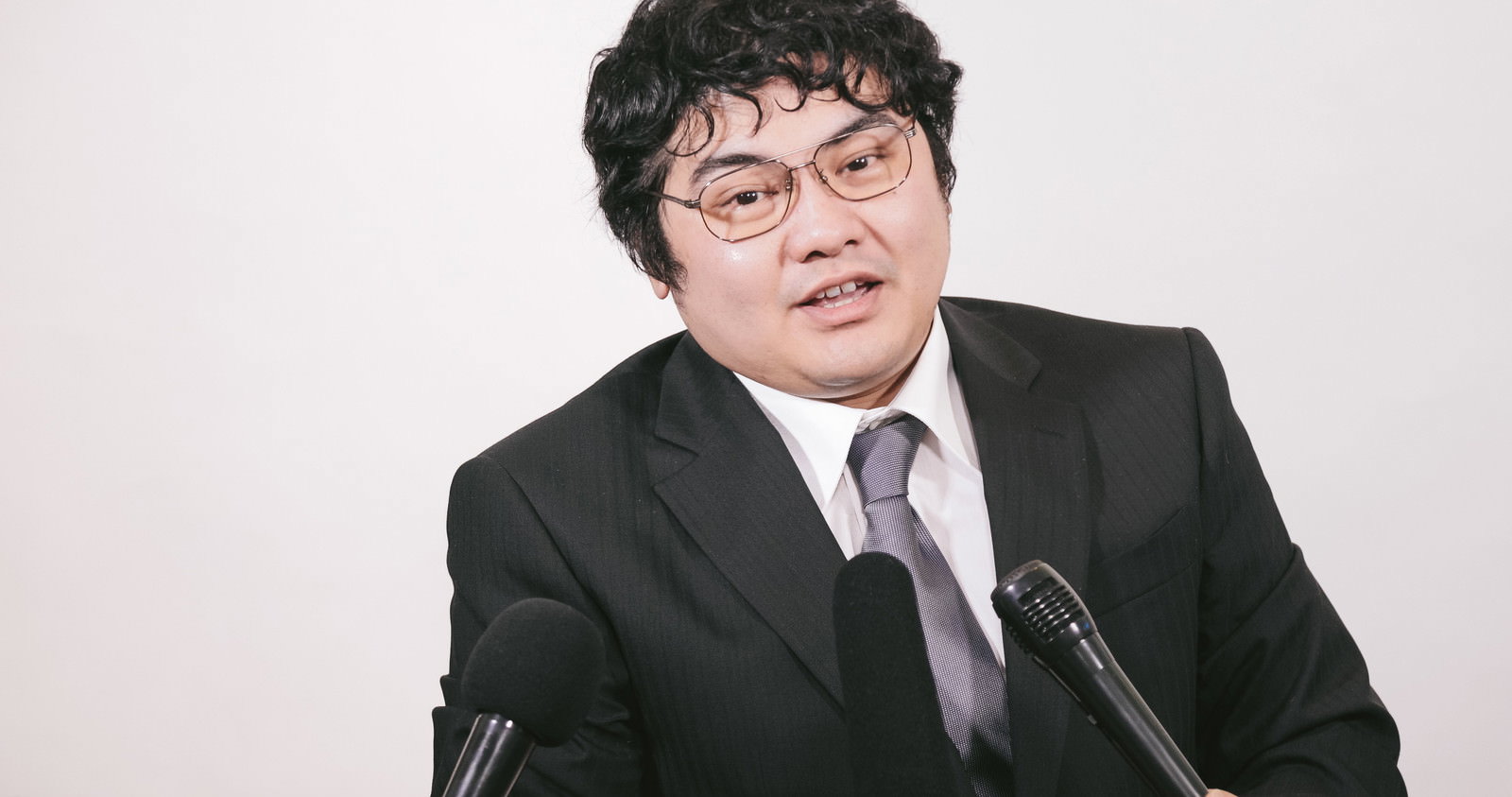 記者会見でマイクに向かって答えるスーツ姿の男性［モデル：段田隼人］