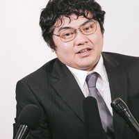 記者会見で複数のマイクに向かって質問に応じるスーツ姿の男性の写真