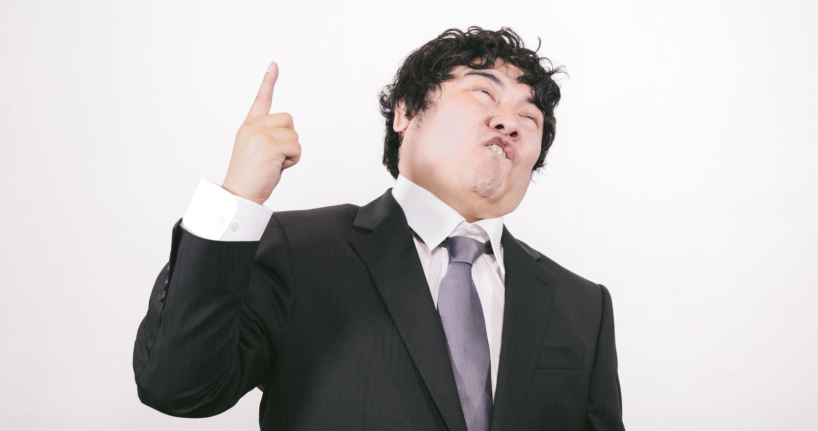 上を指差して主張するスーツ姿の男性［モデル：段田隼人］