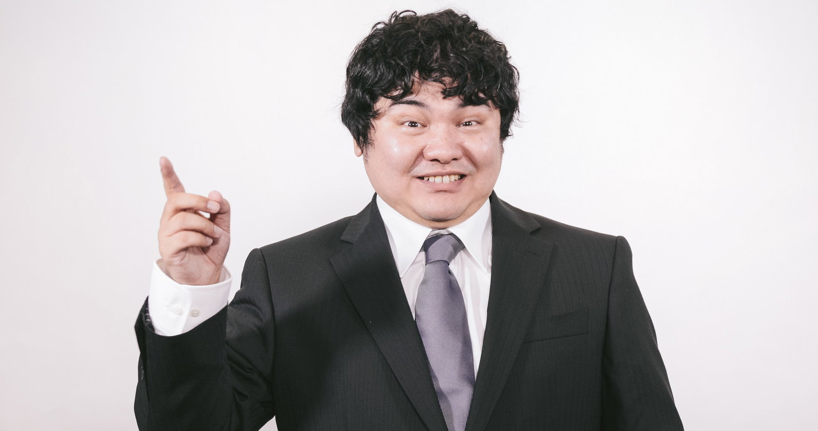 黒いスーツのビジネスマンが右手で上方を指している［モデル：段田隼人］