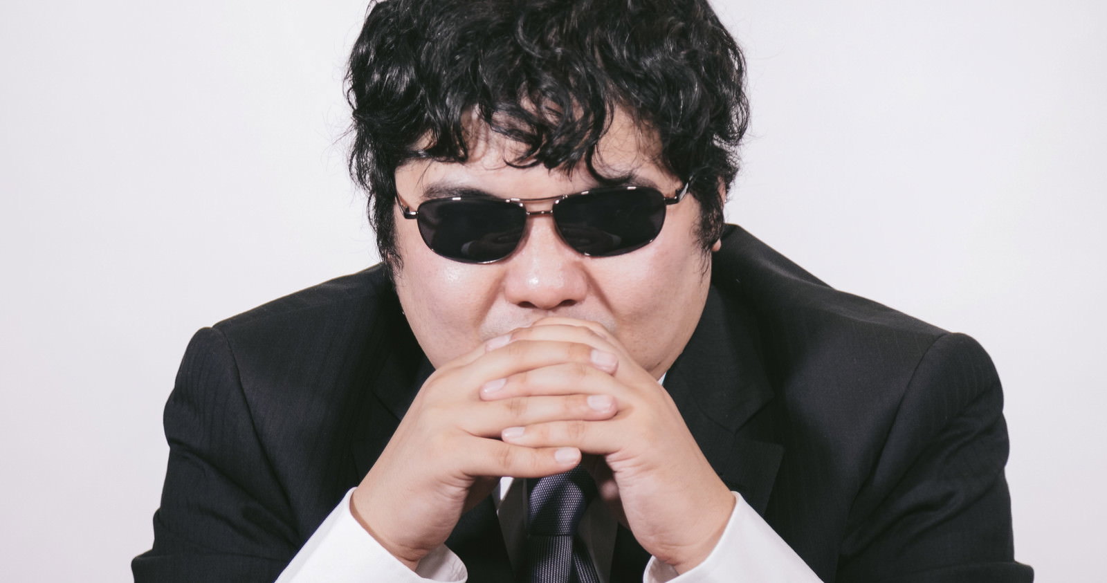 サングラスをかけたスーツの男性が考えるポーズ［モデル：段田隼人］