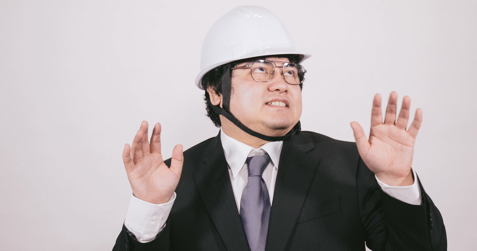 白いヘルメットを被ったスーツ姿の男性が両手を上げている［モデル：段田隼人］