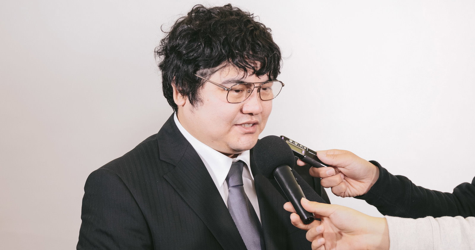複数のマイクを向けられスーツ姿で対応する眼鏡の男性［モデル：段田隼人］