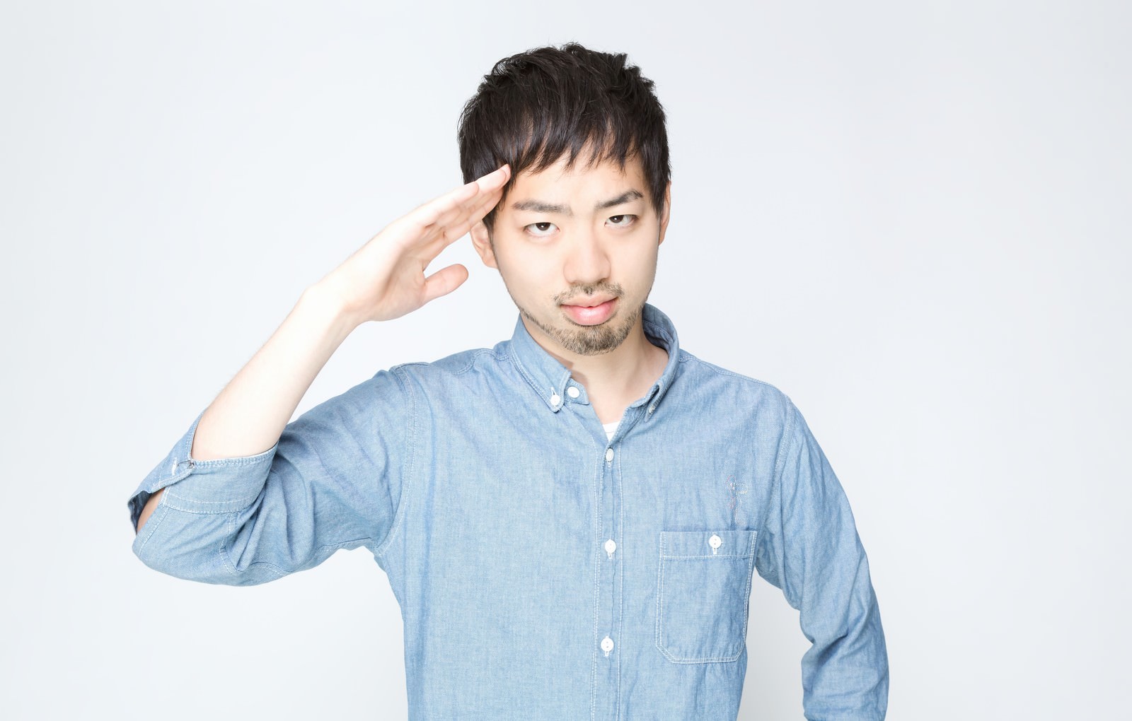 白背景のスタジオで青いシャツの男性が右手で敬礼のポーズをとっている［モデル：ひろゆき］
