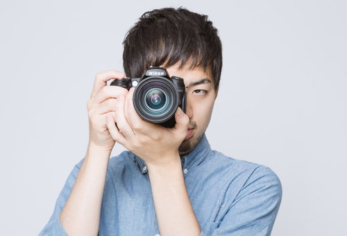 黒いカメラを正面に構える水色シャツの男性
