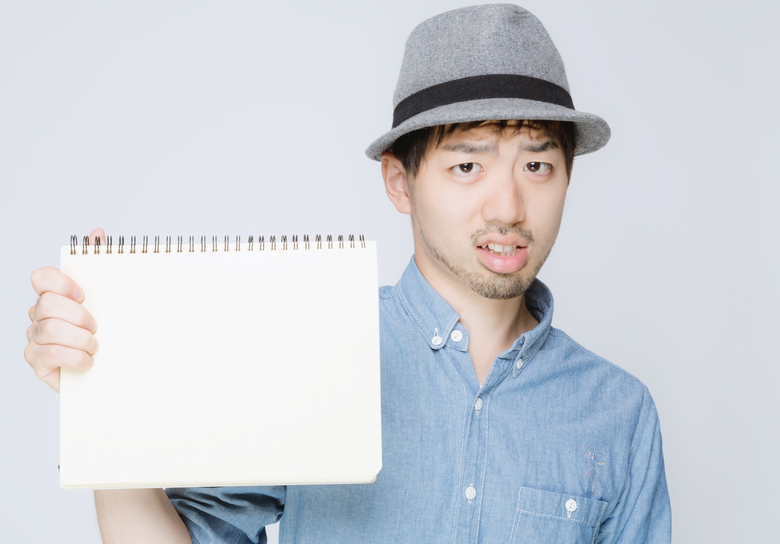 灰色のハットの男性が白い無地のスケッチブックを手に持ち正面を向いている［モデル：ひろゆき］