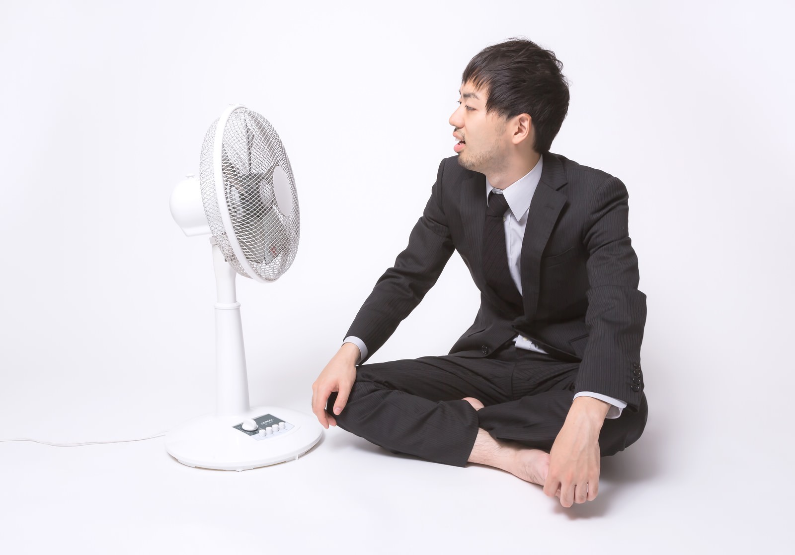 白い扇風機の前で口を開けて涼むスーツのサラリーマン［モデル：ひろゆき］