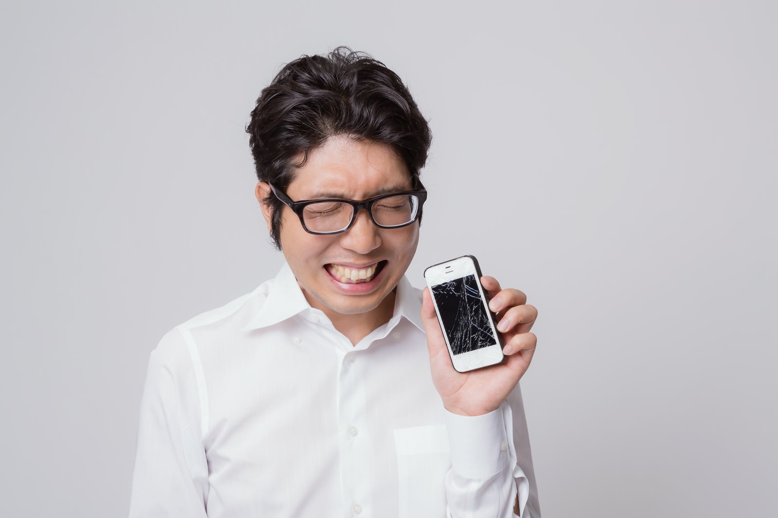 画面の割れたスマートフォンを持ち、悔しそうな表情で見下ろす眼鏡をかけた白シャツの男性［モデル：紳さん］