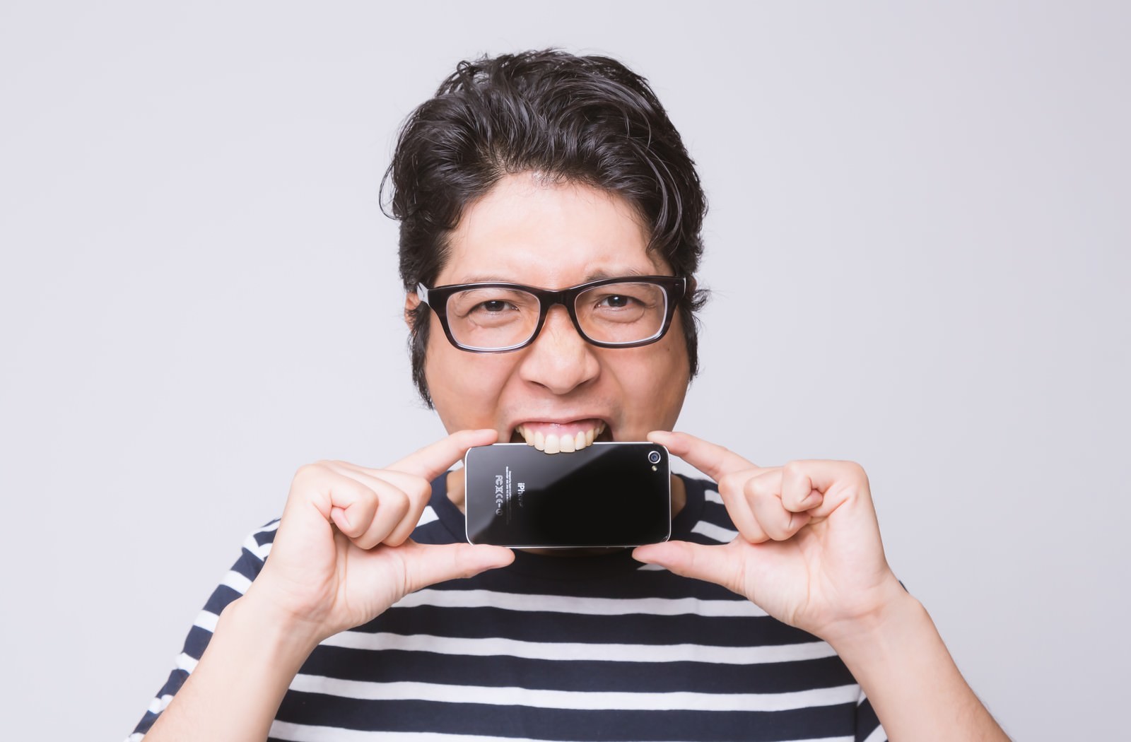 スマートフォンを齧る黒縁眼鏡の男性アプリ開発者［モデル：紳さん］