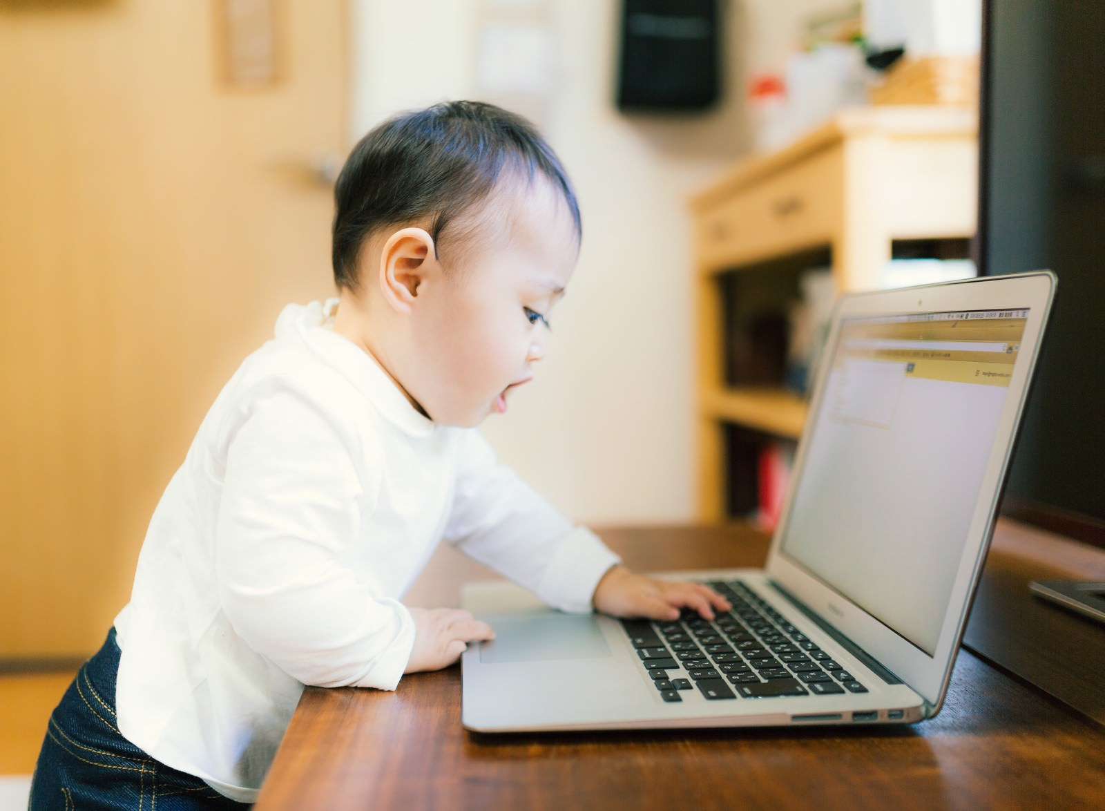 ノートパソコンを前に集中する白い服の乳児、両手でキーボード操作をしようとしている［モデル：Lisa］