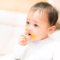 黄色い離乳食のスプーンを口に入れる短い黒髪の赤ちゃんの写真
