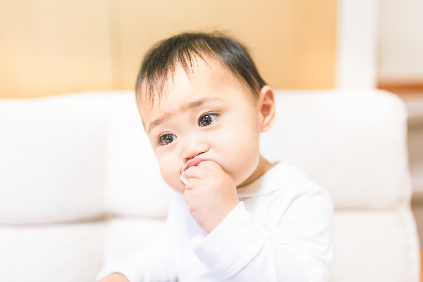 白いソファで手を口元に当てる赤ちゃん［モデル：Lisa］