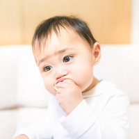 白いソファで手を口元に当てる白い服の赤ちゃんの写真