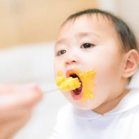 大人の手のスプーンから黄色い離乳食を食べる赤ちゃんの写真