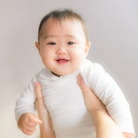 大人の両手に支えられて口を開けて笑う白い服の赤ちゃんの写真
