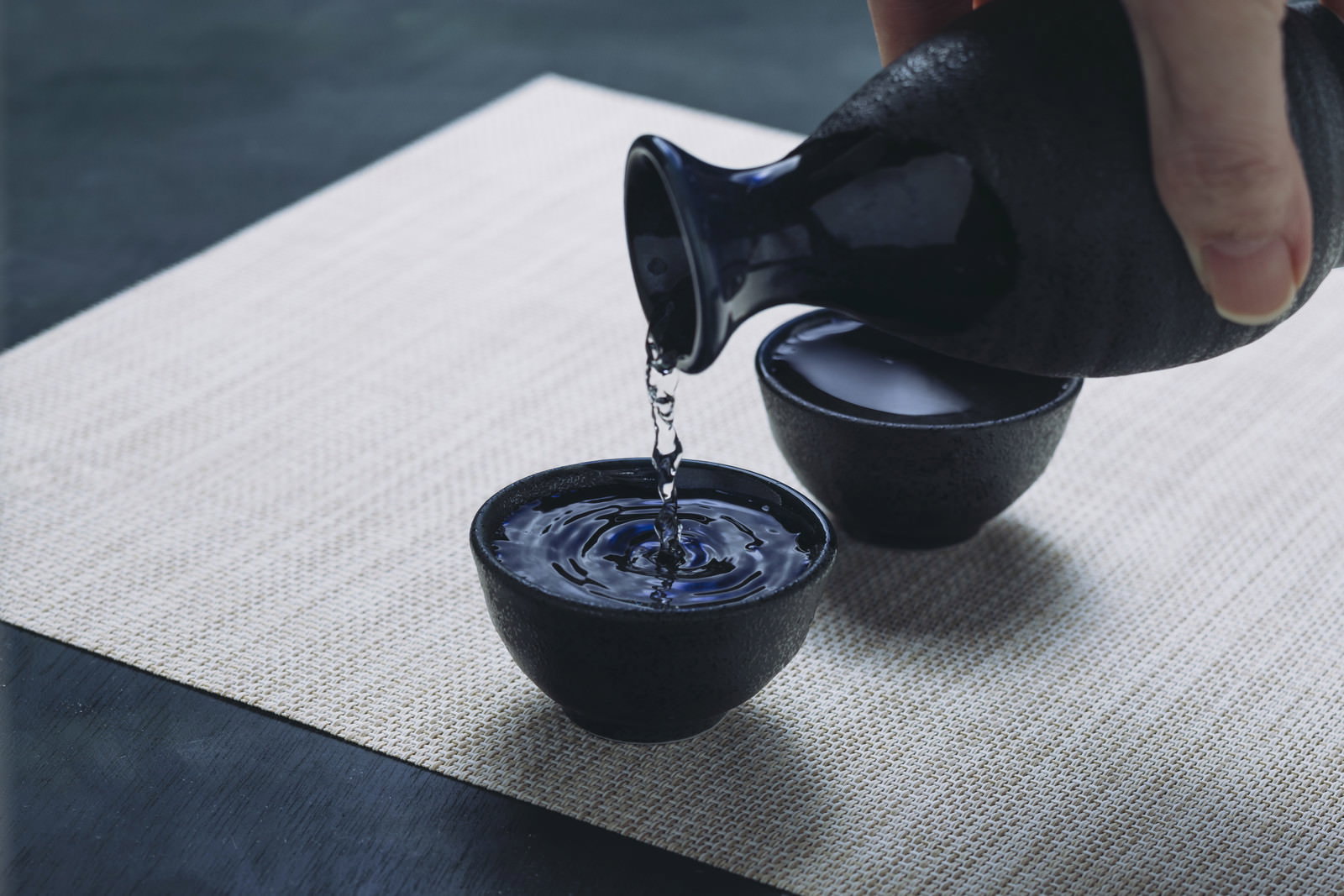畳の上で黒い徳利から青いお猪口に日本酒を注いでいる様子