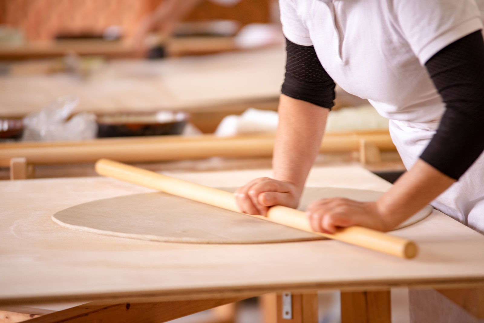 木製のめん棒で蕎麦生地を伸ばす白い作業着の職人