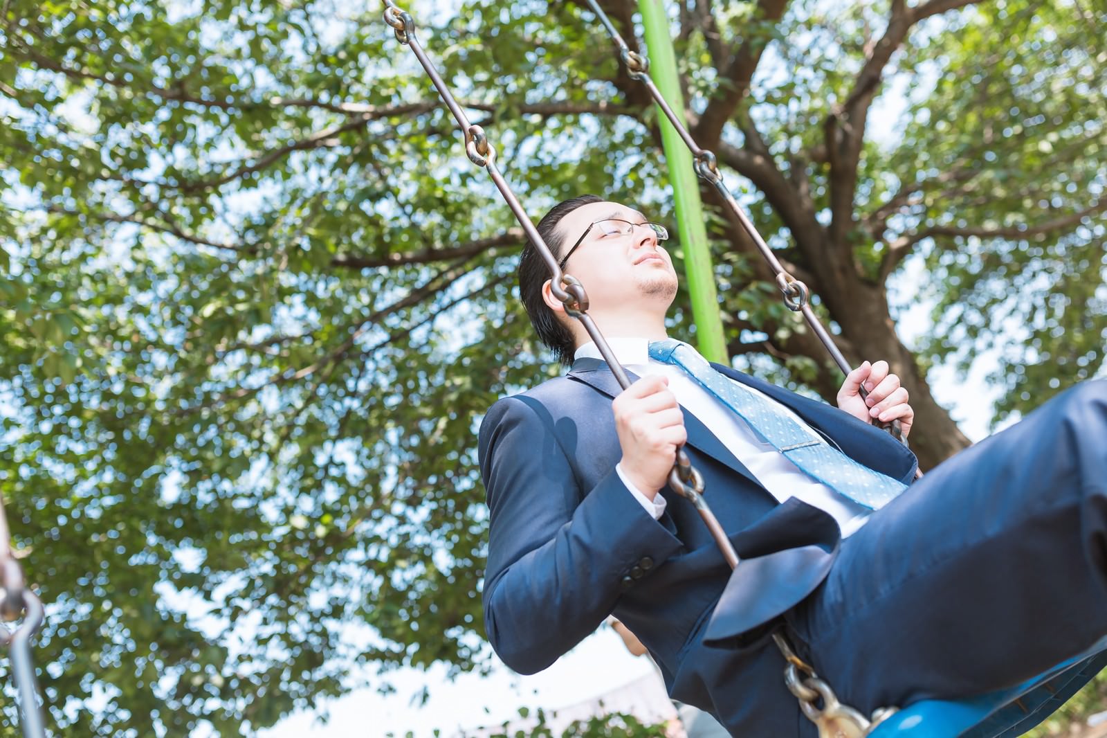 公園のブランコに座って顔を上げるスーツの眼鏡の男性［モデル：Max_Ezaki］