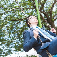 公園のブランコに座って顔を上げるスーツの眼鏡の男性の写真