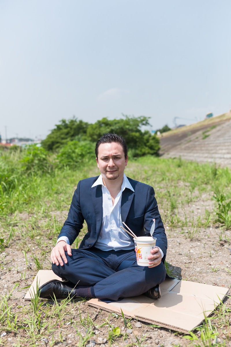 段ボールの上でカップ麺を食べる紺スーツの男性［モデル：Max_Ezaki］