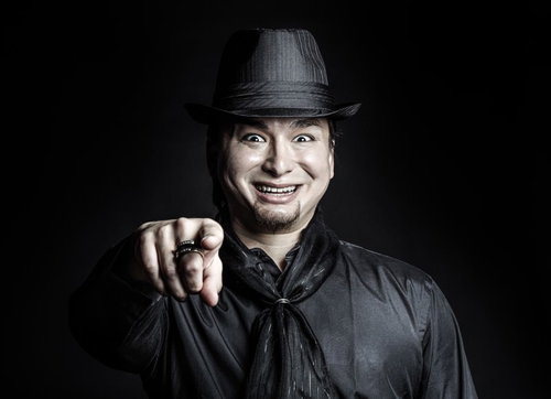 暗い背景で正面を向き笑顔で指を向ける帽子の黒い服の男性