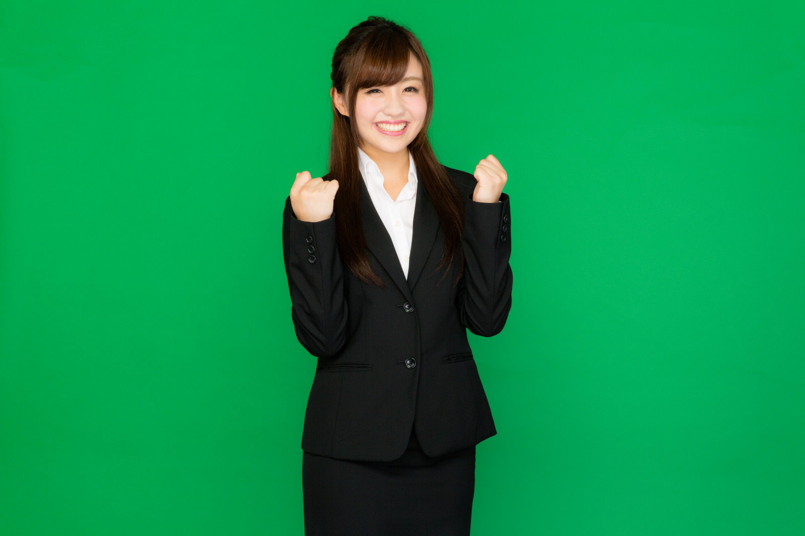 グリーンバックの前で両こぶしを上げてポーズする黒スーツの女性が笑顔で正面を向いている［モデル：河村友歌］