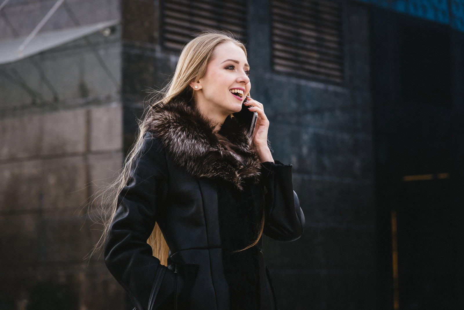 黒いコートを着た外国人女性がスマートフォンで通話しながら笑顔を見せている屋外での様子［モデル：モデルファクトリー］