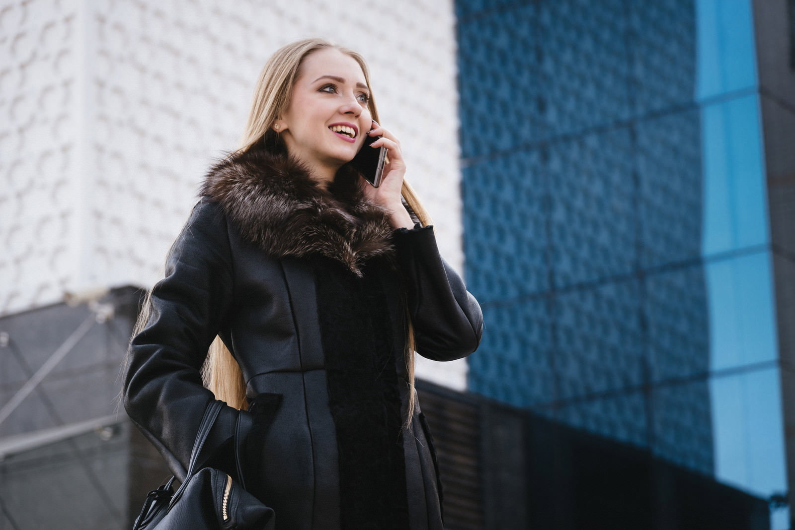 黒いコートを着た金髪の外国人女性がスマートフォンで電話をしている［モデル：モデルファクトリー］